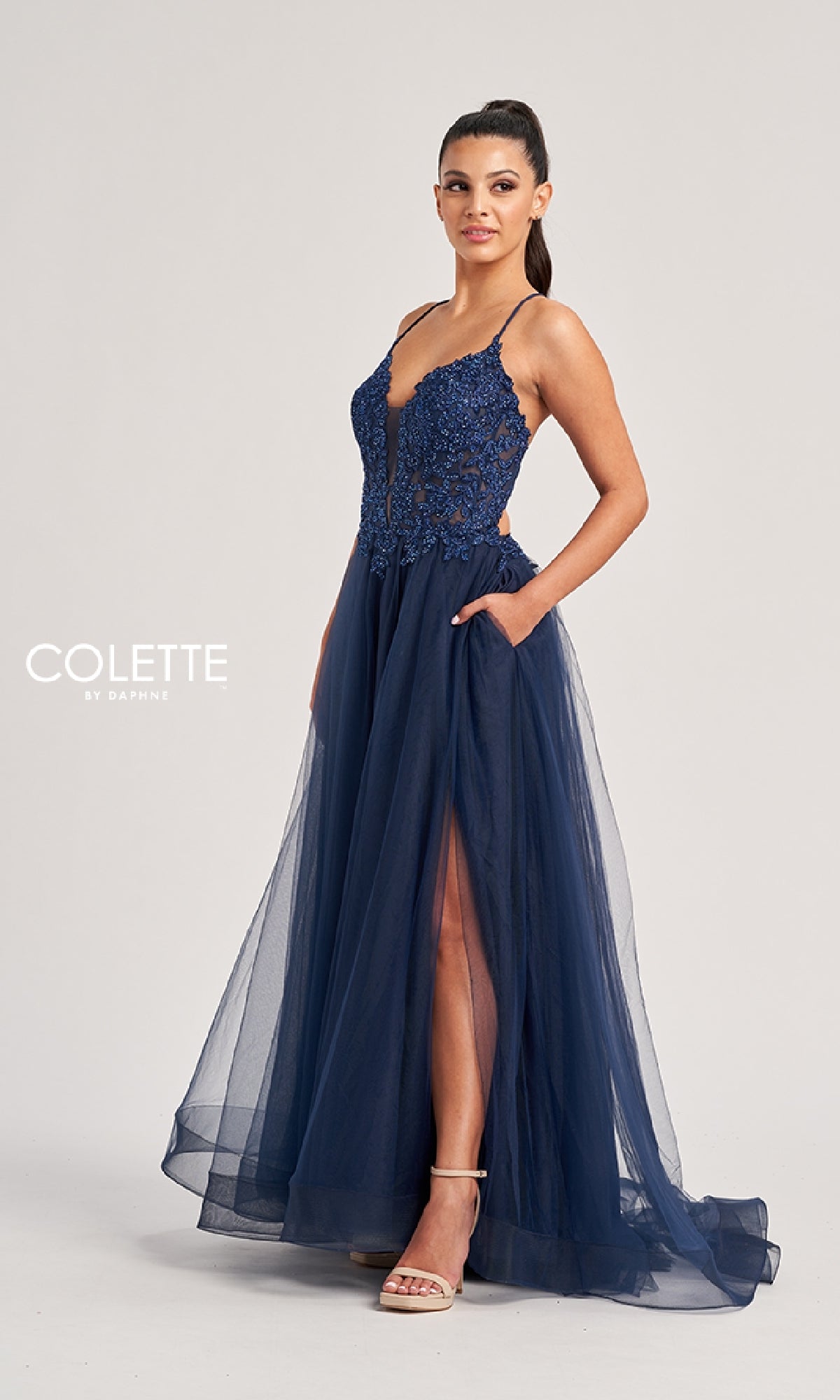A woman stands against a plain background wearing the Colette CL8320, a navy blue long prom dress featuring an embroidered bodice, sleeveless design, spaghetti straps, tulle A-line skirt, and a high slit.